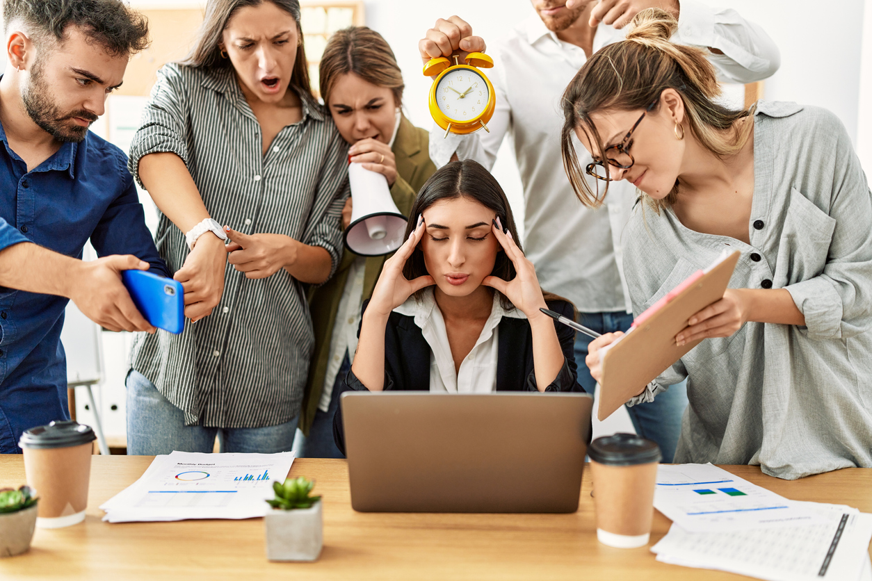 Professional woman at desk is surrounded by demanding coworkers yelling at her and holding clocks, phones, and other signs of deadlines