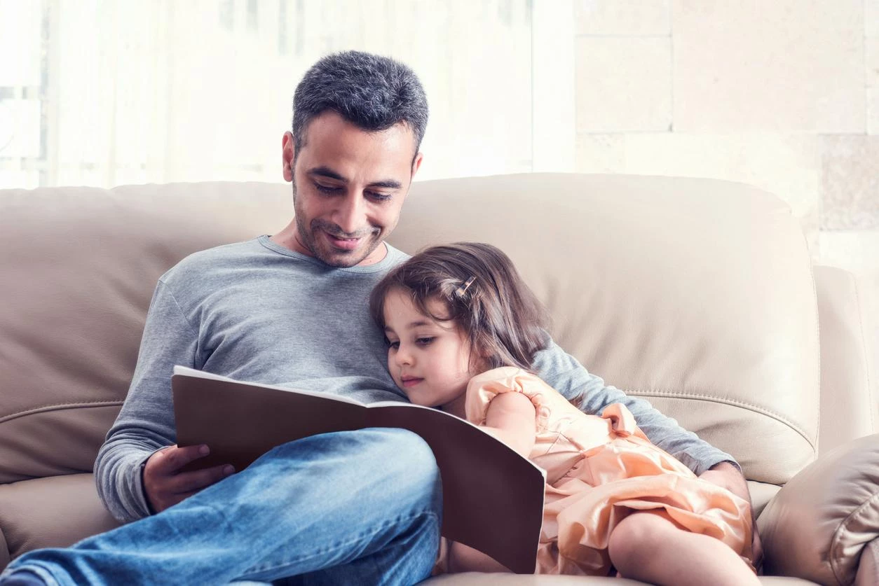 Dad and daughter snuggle on the sofa while reading a book due to disability benefits